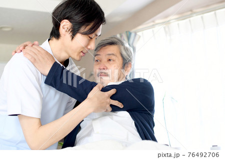 ベッドでお年寄りの男性を起こす介護士の男性 76492706
