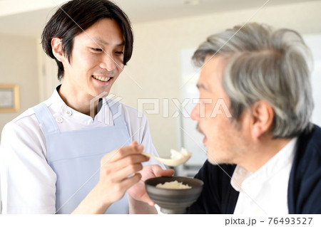 シニア男性に食事の介助をするエプロン姿の若い男性 シニア男性に食事の介助をするエプロン姿の若い男性 76493527