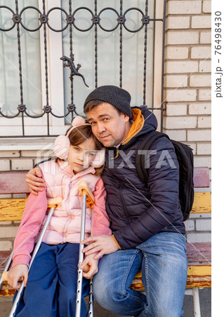 dad with daughter with crutches sits on bench in yard of apartment building 76498430