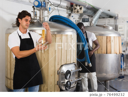 Serious young man brewer attentively checking quality of beer in flask 76499232