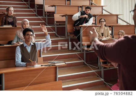 Young successful student in eyeglasses and casualwear raising hand and looking at professor 76504882