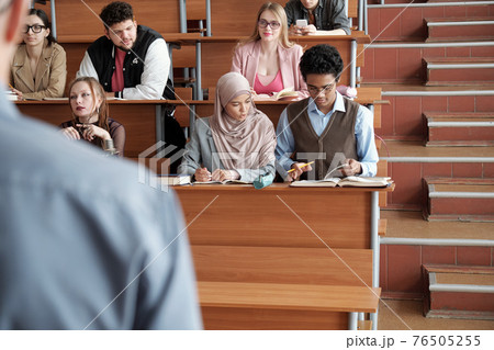 Several rows of contemporary intercultural university students sitting by desks in lecture hall 76505255