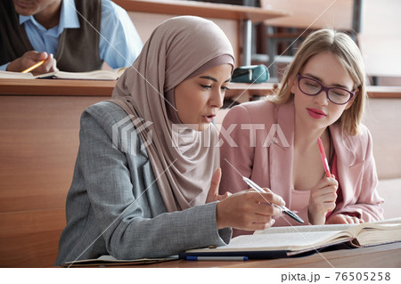 Two intercultural female students sitting by desk in lecture hall and discussing passage in book Two intercultural female students sitting by desk in lecture hall and discussing passage in book 76505258