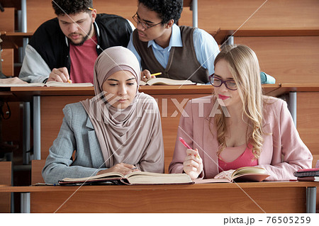 Two intercultural female students of university sitting by desk and looking through passage in book 76505259