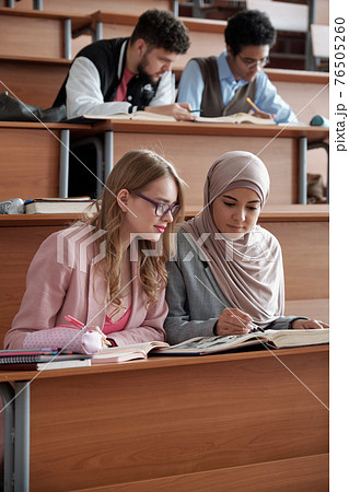 Two intercultural female students of university sitting on the first row by desk in lecture hall Two intercultural female students of university sitting on the first row by desk in lecture hall 76505260