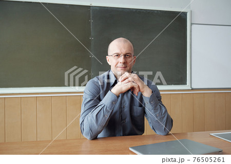 Bald professor of modern university sitting by desk against blackboard while keeping hands by chin 76505261