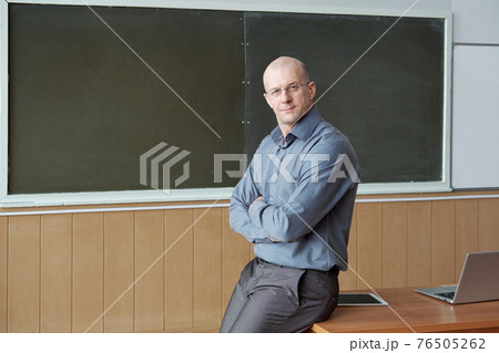 Bald professor of university in smart casualwear sitting on desk against blackboard 76505262