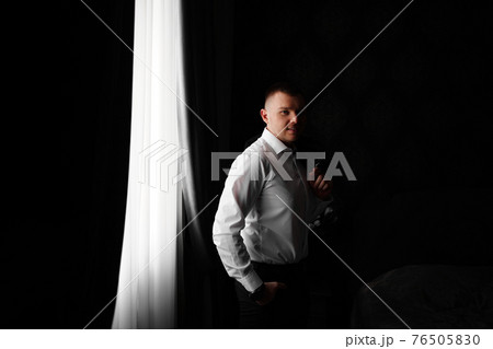 smiling man in a white shirt with a jacket in hands by the window in a dark room 76505830