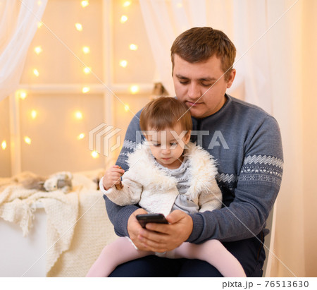 father and baby use smartphone, they play and watch device screen, home interior decorated with holiday lights and gifts 76513630