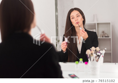 girl, makeup artist, with long dark hair in a business suit sits at the makeup table in a bright room, office and paints his lips girl, makeup artist, with long dark hair in a business suit sits at the makeup table in a bright room, office and paints his lips 76515102