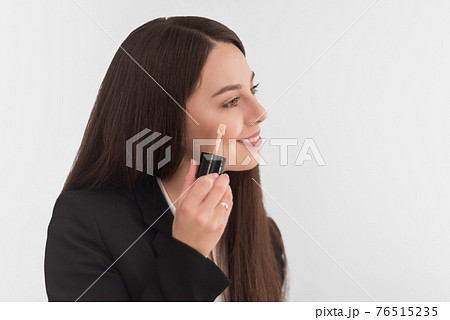 girl, makeup artist, with long dark hair in a business suit, applies a concealer on the face on a white background 76515235