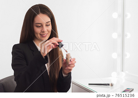 girl, makeup artist, with long dark hair in a business suit sits at the makeup table in a bright room, office and mixes the foundation 76515236