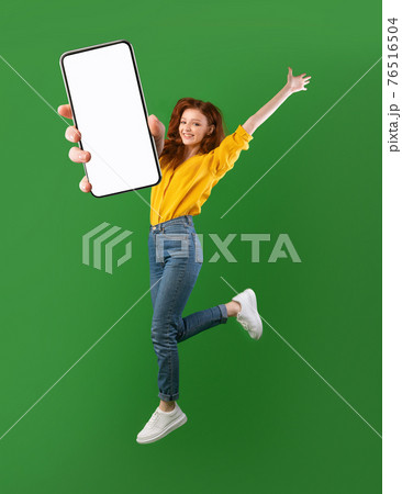 Hot offer, online sale. Carefree young woman jumping in air, showing cellphone with mockup space over green background 76516504