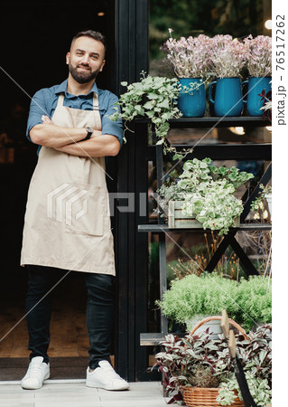 Cheerful millennial confident guy in apron with crossed arms in front door of flower shop Cheerful millennial confident guy in apron with crossed arms in front door of flower shop 76517262