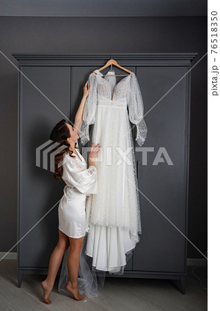 bride in a silk robe next to a white wedding dress hanging on the closet. bride in a silk robe next to a white wedding dress hanging on the closet. 76518350