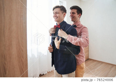 Gay man helping to put on a clothes to his boyfriend while standing with him 76524006