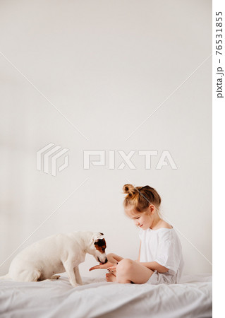 Beautiful funny kid girl feeds with the palm of hand a dog in white bed.  76531855