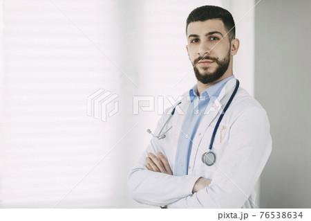 Serious young doctor in stethoscope and white medical coat stands in his office with arms crossed Serious young doctor in stethoscope and white medical coat stands in his office with arms crossed 76538634