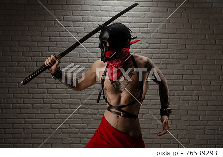a samurai man in a demon mask with tattoos and a katana sword in his hands against the background of a brick wall 76539293