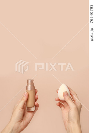 Applying foundation on makeup sponge. Woman's hands with neutral manicure holding bottle of concealer or toner foundation, cream and beauty blender, make up artist background 76540795