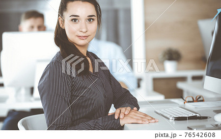 Business woman using computer at workplace in modern office. Brunette secretary or female lawyer looking at the camera and happy smiling. Working for pleasure and success 76542608