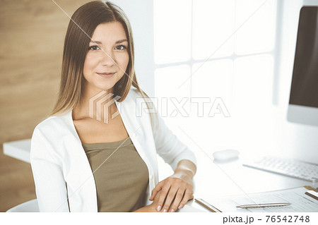 Cheerful smiling businesswoman sitting at the desk in modern office. Young female designer or auditor headshot. Business people concept Cheerful smiling businesswoman sitting at the desk in modern office. Young female designer or auditor headshot. Business people concept 76542748