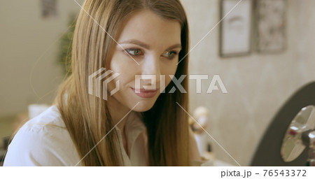 A young woman looks in the mirror, preens herself, admires herself, straightening her clothes and hairstyle. Smiles and likes herself. Start to new day, enjoying morning. Beauty salon makeup 76543372