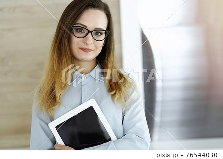 Business woman headshot in sunny office. Unknown businesswoman standing straight with tablet computer. Young accountant or secretary looks good Business woman headshot in sunny office. Unknown businesswoman standing straight with tablet computer. Young accountant or secretary looks good 76544030