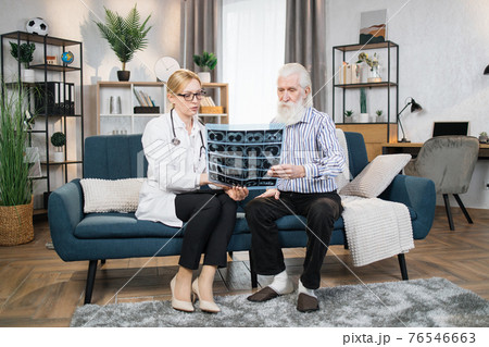 Middle aged attractive high-skilled caring female doctor explaining results of x-ray MRI image to her senior male patient during visiting him at home. Computed tomography 76546663