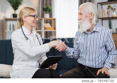 Front view of good-looking professional female Caucasian doctor in medical coat, consulting her senior patient bearded man and handshaking after sighning an insurance, during home visit Front view of good-looking professional female Caucasian doctor in medical coat, consulting her senior patient bearded man and handshaking after sighning an insurance, during home visit 76546730