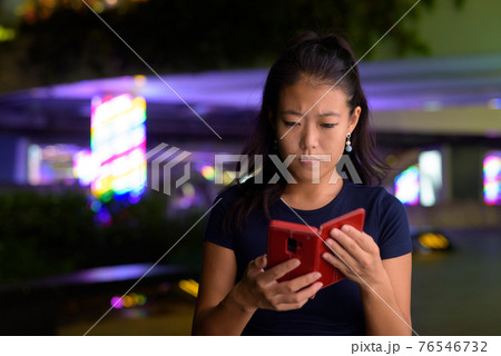 Portrait of beautiful Asian woman outdoors at night using mobile phone 76546732