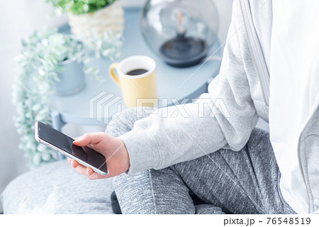 自宅のリビングでスマホを手にする若い男性の手元 76548519