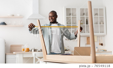 Excited black man measuring furniture with roulette, assembling new table in kitchen interior Excited black man measuring furniture with roulette, assembling new table in kitchen interior 76549118