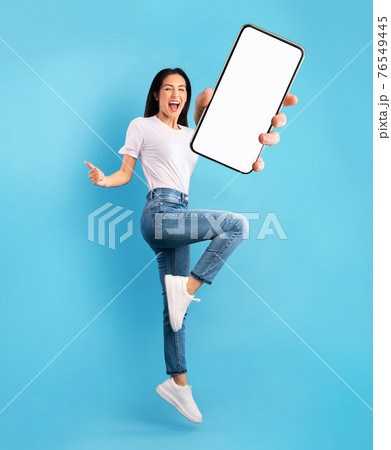 Advertisement for mobile app. Excited lady jumping over blue background, showing modern smartphone with empty screen 76549445