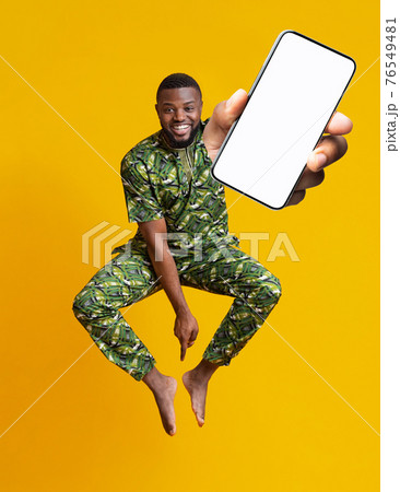 Barefoot happy black guy in African costume jumping up with smartphone over orange studio background, mockup 76549481