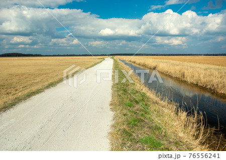 Gravel road and water canal next to the meadows, clouds on the sky 76556241