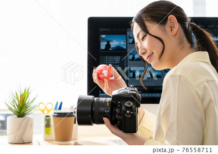 PCの前でデジタル一眼レフカメラをメンテナンスする女性クリエイター 76558711
