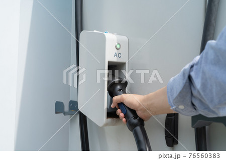 Hand pulling in cable from EV car charger or electric vehicle station. Cable connected at gas station, power supply battery charging an alternative eco environment future technology energy innovation 76560383