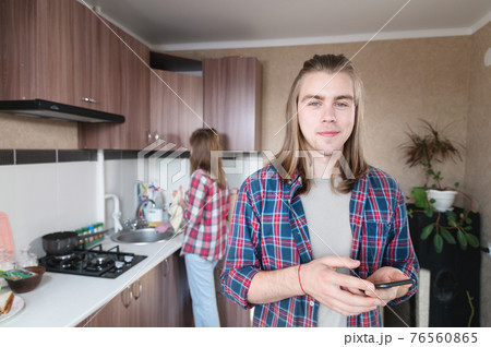 in the kitchen room, a young man with long hair stands with a phone in his hands and looks at the camera 76560865