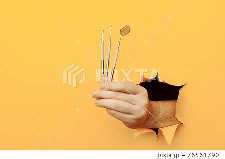 Hand showing a dentist tools out of a hole torn in yellow wall. Health care concept 76561790
