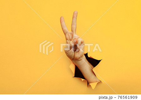 Hand showing peace gesture through torn yellow paper background. Victory, V sign 76561909
