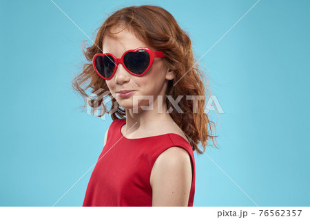 Little girl in sunglasses in the form of hearts red dress curly hair blue background 76562357