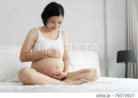 Cheerful Asian Young pregnant woman smiling sitting touching her beautiful belly on bed at home. Cheerful Asian Young pregnant woman smiling sitting touching her beautiful belly on bed at home. 76573627