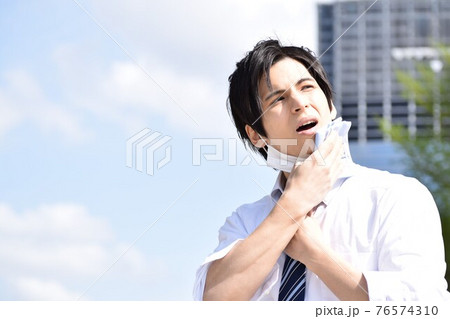 暑くてマスクを外してハンカチで汗を拭いている若い男性 76574310