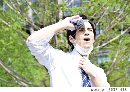 暑くてマスクを外してハンカチで汗を拭いている若い男性 76574458