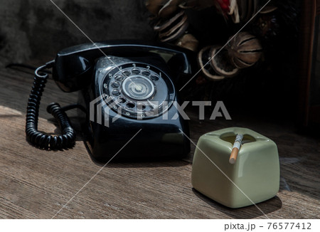 A litted cigarette on greyish green ashtray which is placed next to old-fashion telephone. The heart-shape wreath as background. A litted cigarette on greyish green ashtray which is placed next to old-fashion telephone. The heart-shape wreath as background. 76577412