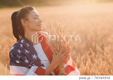 Woman with American flag and with a sheaf of ears in wheat field at sunset. 4th of July. Independence Day patriotic holiday. 76584950