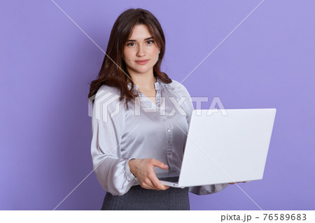 Young woman with dark hair using laptop and smiling isolated over lilac background, looking at camera, wearing elegant blouse and skirt, beautiful female with portable computer indoor. 76589683