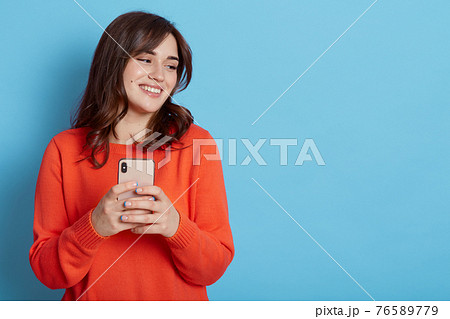 Carefree pleased female standing with happy look and holding mobile phone, looks aside and smiles broadly, browses through internet during free time, posing indoor, dresses casual attire. 76589779