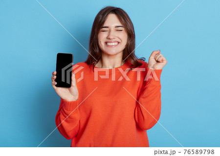 Overjoyed attractive woman keeps fist clenched, being happy to get good news, gets notification on cellphone, wears orange sweater, stands with closed eyes indoor against blue wall, feels amused. 76589798